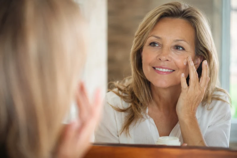 Smiling woman applying a compounded topical skincare formulation in the mirror
